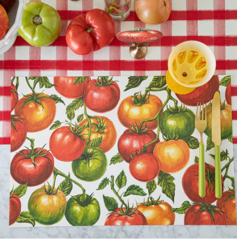 A table features a red-and-white checkered cloth, tomatoes, a yellow citrus juicer, gold utensils, and the Hester & Cook Tomato Vine Placemat - Pad of 18 Sheets with tomato illustrations in red, green, and yellow shades with leafy stems.