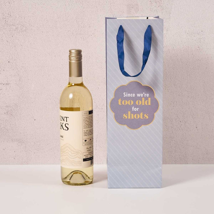 A bottle of white wine sits beside the Drinks On Me "Too Old- Wine Bag," a light blue reusable bag with dark blue handles and a gold-edged label that reads, "Since we're too old for shots.
