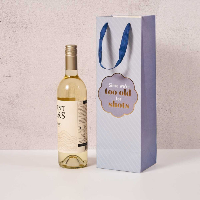 A bottle of white wine sits beside the Drinks On Me "Too Old- Wine Bag," a light blue bag with a ribbon handle and gold text reading, "Since we’re too old for shots.