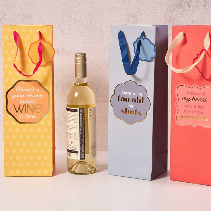 Three colorful Drinks On Me "Too Old" wine bags in yellow, blue, and red stand beside a bottle of white wine. Each reusable bag features a humorous text and ribbon handle, making them fun gift options.