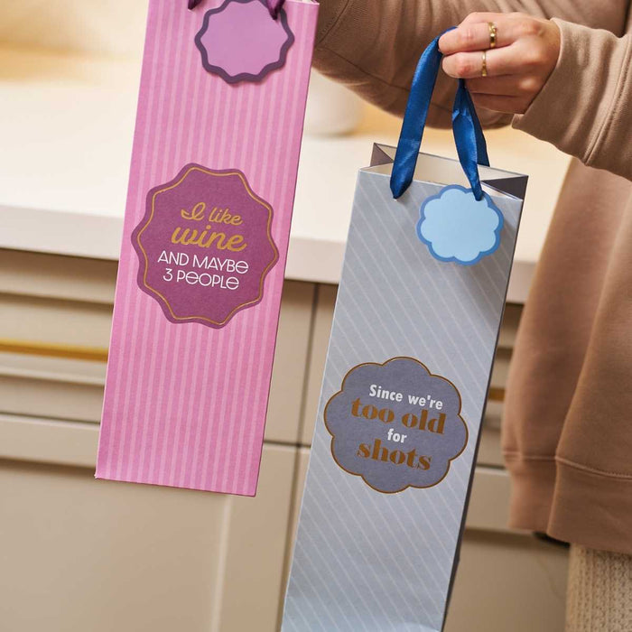 Drinks On Me's Too Old-Wine Bag set features two reusable gift bags: one pink with "I like wine and maybe 3 people," and one blue with "Since we're too old for shots." Each bag fits a standard wine bottle perfectly.