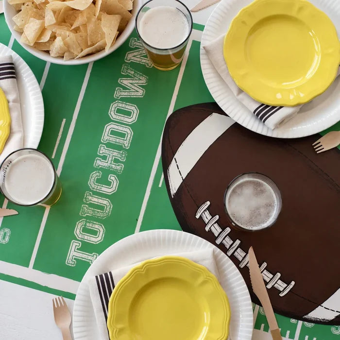 A football-themed party table features yellow plates, striped napkins, wooden cutlery, beer glasses, tortilla chips, and a Hester & Cook Touchdown Runner (20x25") styled like a football field with TOUCHDOWN text and a large football.