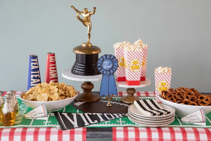 A football-themed party table with snacks, drinks, plates, and napkins features festive decor—trophy, ribbon, pennants, megaphones—and a Hester & Cook Touchdown Runner (20x25") as a standout centerpiece.