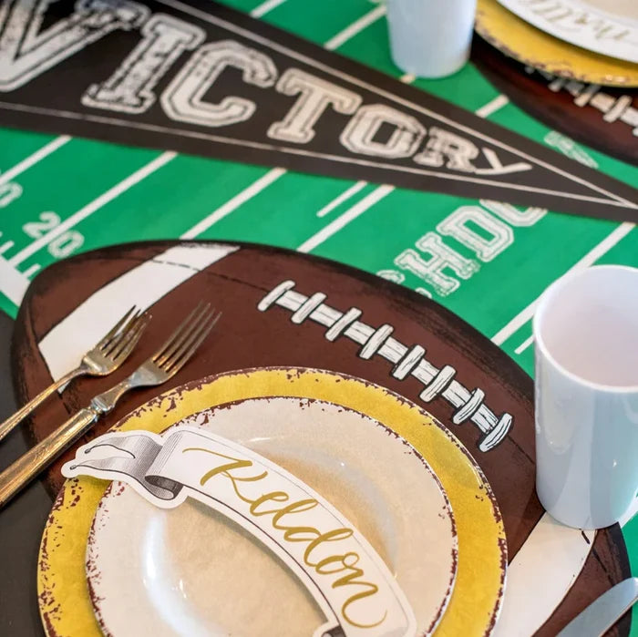 A football-themed table is set with a brown and white football placemat, gold-rimmed plate, utensils, white cup, name card for Keldon, Victory banner, and the green Touchdown Runner (20x25") by Hester & Cook as a paper table runner.