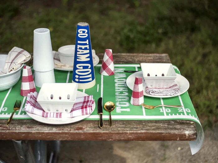 A picnic table outdoors on grass is decorated with the Hester & Cook Touchdown Runner (20x25"), football field-themed plates, cups, utensils, napkins, and a blue “GO TEAM GO!” megaphone.