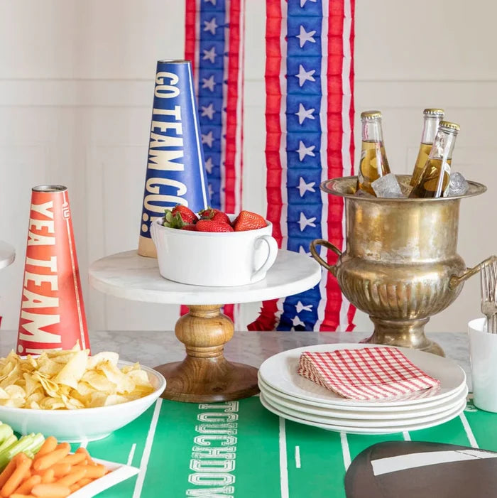 A football-themed party table features snacks, drinks on ice, strawberries, chips, veggies, megaphones, and patriotic star streamers—all displayed atop the Hester & Cook Touchdown Runner (20x25") for a festive touch.