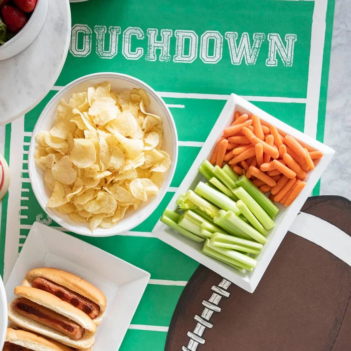 A tabletop features the Hester & Cook Touchdown Runner (20x25") with a green football field design, displaying bowls of potato chips, celery sticks, baby carrots, hot dogs, and a football-shaped plate.