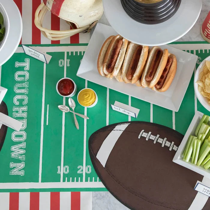 A football-themed table features a field-patterned cloth, Hester & Cook's Touchdown Runner (20x25"), hot dogs, chips, celery sticks, cups of ketchup and mustard, football-shaped placemats, and a partially visible football helmet.