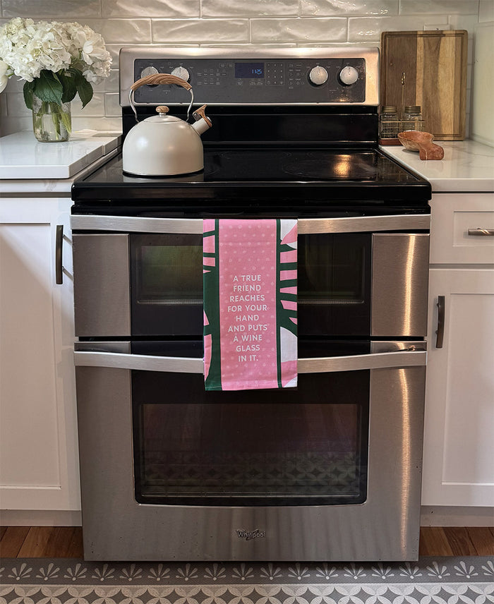 A modern stainless steel oven with a white kettle on top and the "True Friend Dish Towel" by Drinks On Me, featuring the quote "A true friend reaches for your hand and puts a wine glass in it." White cabinets and flowers nearby.