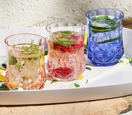 Three Sophistiplate Tumbler Classic Short cups in Blush (15 oz), BPA-free and shatterproof, are filled with fruit and herb-infused drinks—lime, raspberries, mint, and cucumber—displayed on a white tray with colorful patterned designs.