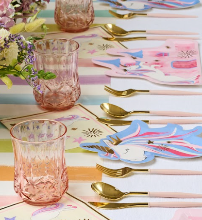A pastel-themed table set with Sophistiplate Tumbler Classic Short Blush 15 Oz cups, gold cutlery, unicorn plates, and floral centerpieces on a softly striped tablecloth.