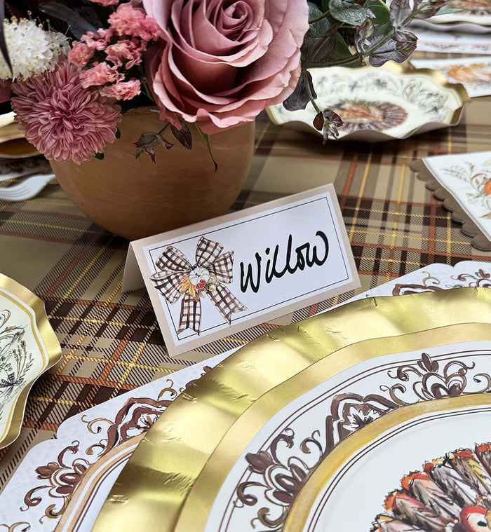 A Rosanne Beck Turkey & Gourds Bow Place Card rests on a plaid tablecloth beside a gold-and-white plate, accented by a checkered bow and surrounded by pink and mauve flowers—perfect for buffet labels or seating.