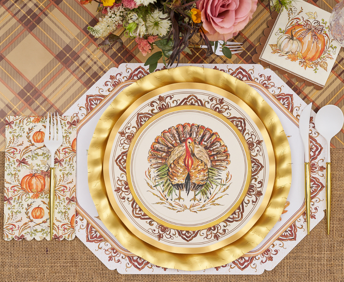 A festive table features Sophistiplate Turkey & Gourds Dinner Plates (8pk), gold-rimmed disposable plates, white cutlery, autumn napkins with pumpkins and florals, all set on a plaid tablecloth with a floral arrangement nearby.