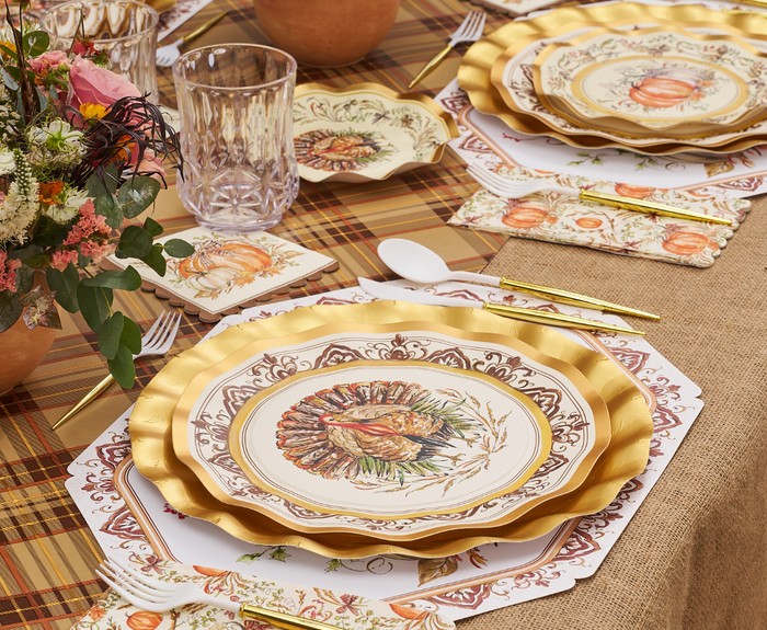 A festive table set with Sophistiplate Turkey & Gourds Dinner Plates (8pk), gold cutlery, glasses, autumn napkins and placemats, plus a fall floral centerpiece on a plaid and burlap tablecloth.