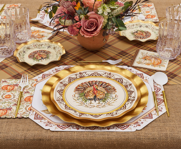 Create an inviting holiday table with the Turkey & Gourds Table Setting Bundle by Sophistiplate LLC, featuring designer disposable plates adorned with a turkey and gold accents, autumn pumpkin-themed napkins, clear glasses, white cutlery, and a floral centerpiece displayed on a plaid and burlap tablecloth.