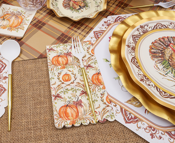 An autumn table setting featuring Sophistiplate's Turkey & Gourds Guest Towels (20pk), plates with a turkey design, gold-edged chargers, white plastic cutlery with gold handles, all set on a plaid and burlap tablecloth.