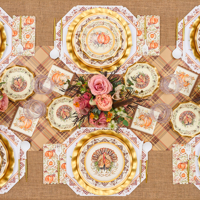 A festive table is set with Rosanne Beck Turkey & Gourds Placemats, autumn plates, gold-rimmed chargers, floral centerpieces with pink roses, fall napkins, and a burlap tablecloth.