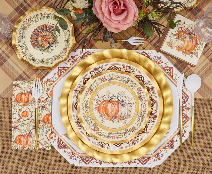 A festive autumn table with gold-rimmed pumpkin plates, a turkey plate, white cutlery, and a centerpiece of pink roses—featuring Sophistiplate’s Turkey & Gourds Cocktail Napkins (26pk) for a charming touch.