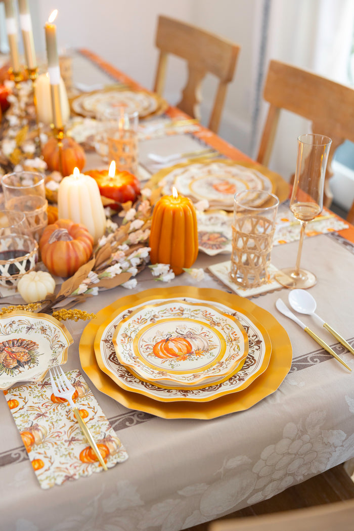Create a festive and cozy atmosphere this autumn with the Sophistiplate LLC Turkey & Gourds Table Setting Bundle, featuring designer disposable plates adorned with seasonal motifs, complemented by candles, miniature pumpkins, gold flatware, and floral accents on a light tablecloth—an elegant solution for stylish entertaining.