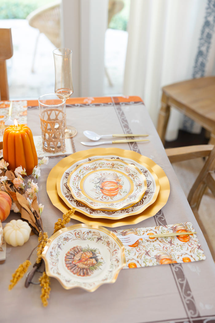 Create a refined autumn table with the Turkey & Gourds Table Setting Bundle from Sophistiplate LLC, featuring designer disposable plates adorned with festive turkey and gourd motifs. This bundle includes gold utensils, a floral napkin, a candle, and charming autumn décor such as a small orange pumpkin and dried leaves on a beige tablecloth. Ideal for effortless entertaining, these heavy-duty paper plates offer both elegance and convenience for your seasonal gatherings.