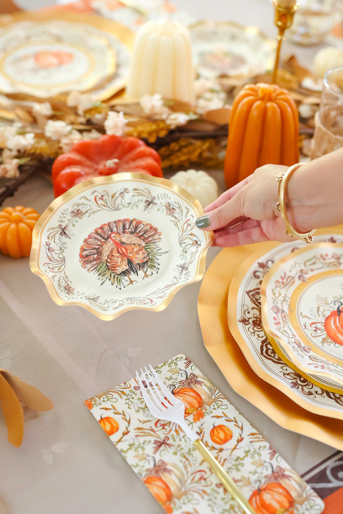 A hand holds a Sophistiplate LLC Turkey & Gourds Table Setting Bundle designer disposable plate with a turkey design over a festive table adorned with coordinating autumn-themed plates, pumpkins, candles, and patterned napkins.