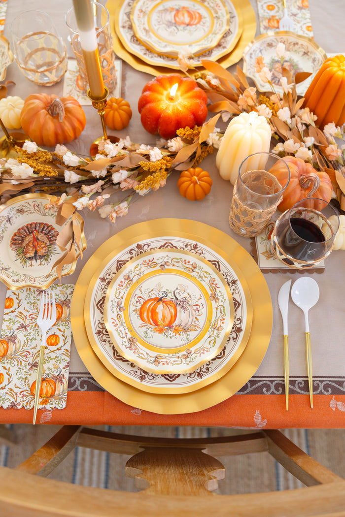 Create a festive Thanksgiving table with the Sophistiplate LLC Turkey & Gourds Table Setting Bundle, which includes designer disposable plates with a pumpkin motif, gold utensils, glassware, a pumpkin-shaped candle, and autumn-inspired decorations such as leaves and mini pumpkins.