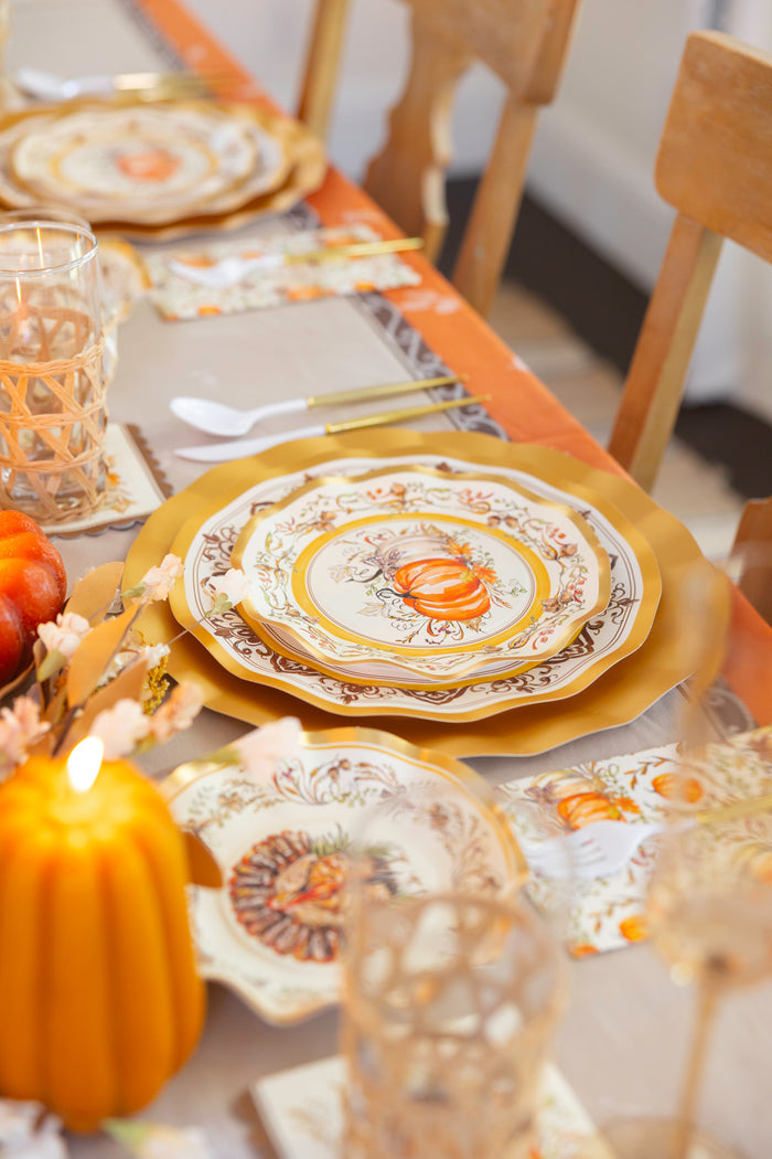 Create a warm and festive atmosphere with the Turkey & Gourds Table Setting Bundle by Sophistiplate LLC, featuring designer disposable plates in a gold finish, pumpkin-printed dishes, elegant gold cutlery, glassware, and a lit pumpkin-shaped candle for an elegant autumn-themed table setting.