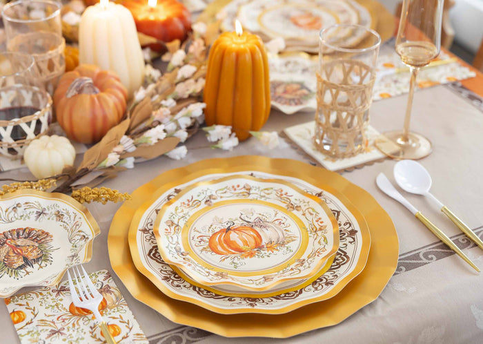 Create a warm and festive atmosphere with the Turkey & Gourds Table Setting Bundle by Sophistiplate LLC, featuring designer disposable plates adorned with gold rims and charming pumpkin motifs, complemented by gold cutlery, clear glasses, candles, faux leaves, and decorative pumpkins—perfect for an elegant autumn-themed table setting.