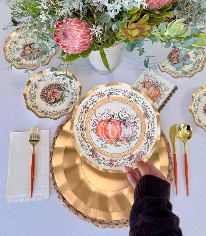 A hand sets the Sophistiplate Turkey & Gourds Table Setting, featuring a designer paper plate, at a fall-themed table with matching napkin, cutlery, and a floral and succulent centerpiece.