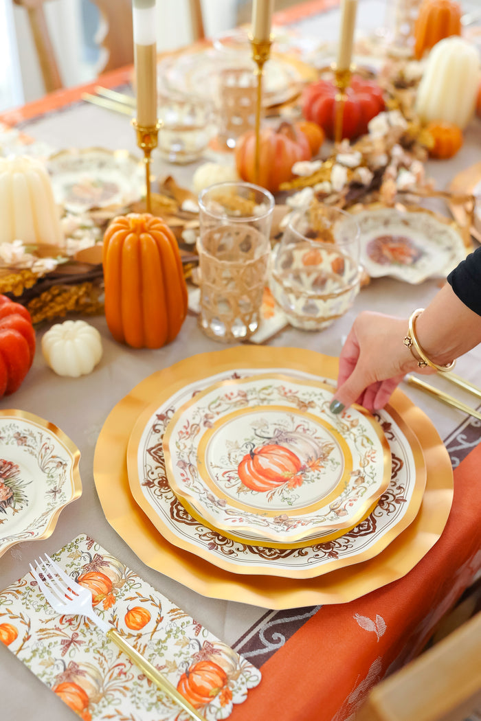 A hand arranges the Sophistiplate Turkey & Gourds Table Setting on an autumn table with Thanksgiving tableware—gold-rimmed plates, candles, glasses, mini pumpkins—plus a fall-patterned cloth and napkin.