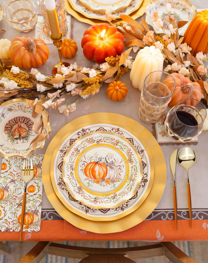Experience a festive autumn table setting featuring the Sophistiplate LLC Turkey & Gourds Table Setting Bundle, complete with pumpkin-themed designer disposable plates, gold utensils, glasses of water and wine, decorative pumpkins, candles, and a fall garland centerpiece arranged on a beige and orange tablecloth.
