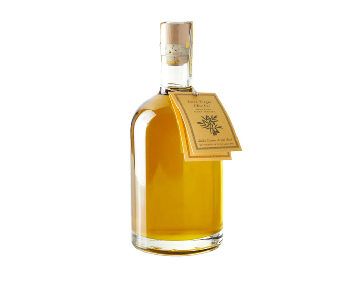 A clear glass bottle of Bella Cucina Tuscany Extra Virgin Olive Oil, filled with golden olive oil and sealed with a cork, features a tan label wrapped around the neck.
