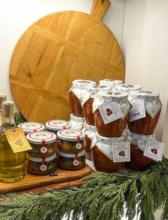 The Bella Cucina Pizza Party Gift Kit—featuring jars of preserves, olive oil, and Italian tomato pizza sauce—is displayed on a counter with greenery, while a large round wooden cutting board stands upright behind it.