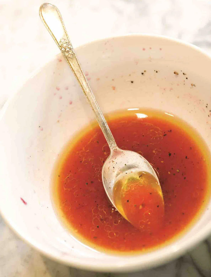 A white bowl holds a reddish vinaigrette made with Bella Cucina Taste of Tuscany Extra Virgin Olive Oil, speckled with black pepper and oil droplets. A silver spoon rests inside the bowl, which sits on a light-colored surface.