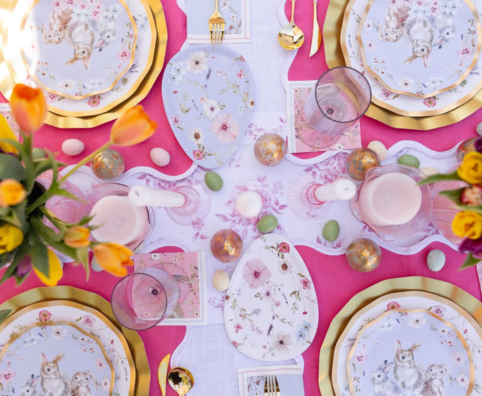 Create a festive spring setting with Sophistiplate LLC’s Ultimate Easter Tablescape for 16: Charming Easter—includes floral and bunny plates, gold cutlery, pink napkins, tulip arrangements, pastel eggs, candles, and glasses on a white-pink cloth.