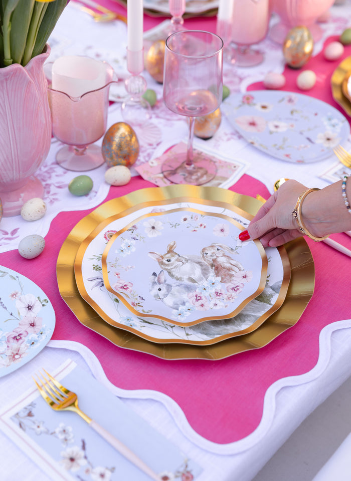 A hand sets a Sophistiplate LLC Ultimate Easter Tablescape for 16: Charming Easter designer plate, featuring rabbit and flower art, onto a gold charger. The festive table includes pastel dishware, pink and white linens, candles, and eggs.