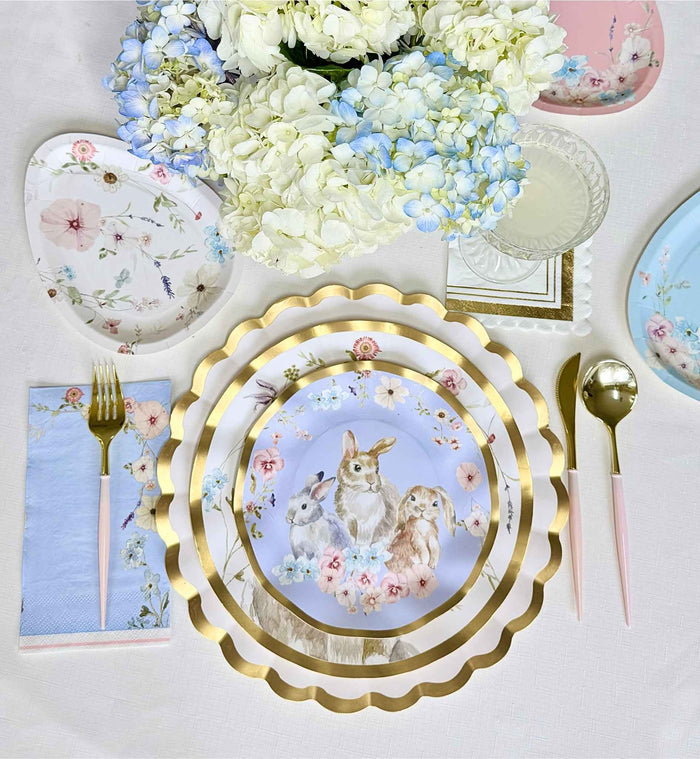 Create a charming spring table with Sophistiplate LLC's Ultimate Easter Tablescape for 16, featuring gold-rimmed scalloped plates with rabbits, pastel napkins, gold and pink cutlery, floral accents, and white and blue hydrangeas.