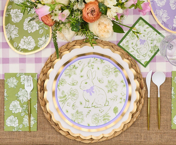 The Sophistiplate LLC Ultimate Easter Tablescape for 16: Easter Toile includes a floral centerpiece, designer bunny-and-flower plates, gold utensils, and floral napkins arranged on a purple gingham tablecloth for spring.