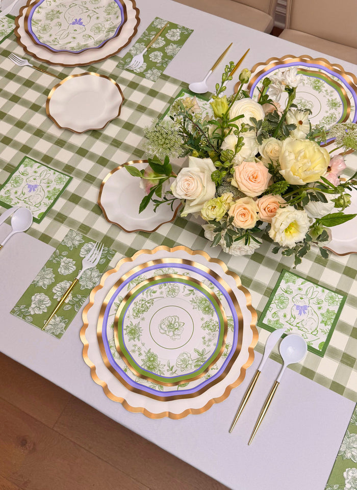 The Ultimate Easter Tablescape for 16: Easter Toile by Sophistiplate LLC features designer plates, gold-rimmed chargers, floral napkins, a green check runner, white cutlery, and a peach-and-white floral centerpiece for a charming holiday setting.