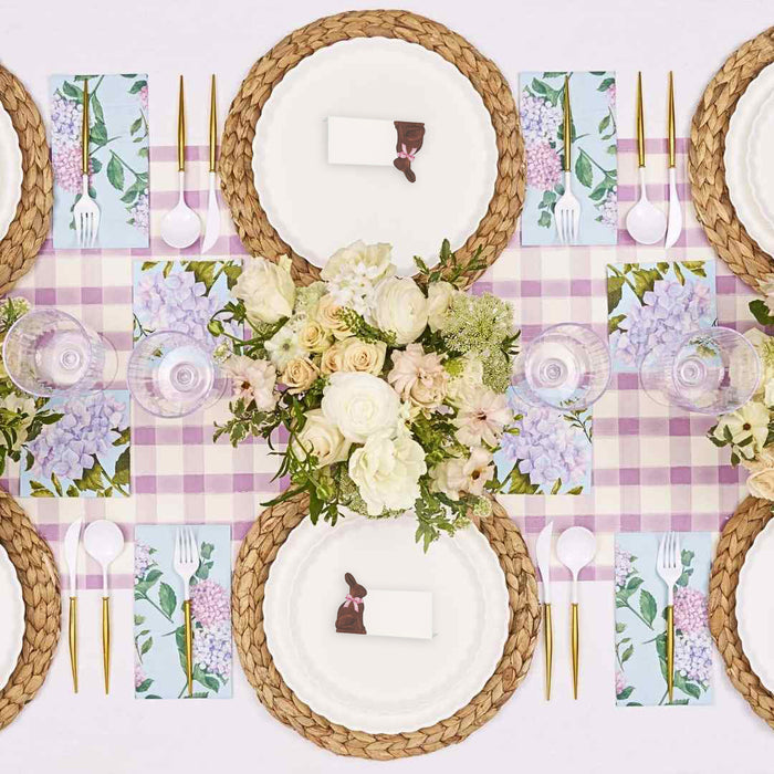 Create a spring-inspired table with the Ultimate Easter Tablescape for 16: Violet Hydrangea by Sophistiplate LLC, featuring woven placemats, designer plates, gold cutlery, floral napkins, name cards with chocolate bunnies, and a white and pink floral centerpiece.