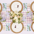 Create a spring-inspired table with the Ultimate Easter Tablescape for 16: Violet Hydrangea by Sophistiplate LLC, featuring woven placemats, designer plates, gold cutlery, floral napkins, name cards with chocolate bunnies, and a white and pink floral centerpiece.
