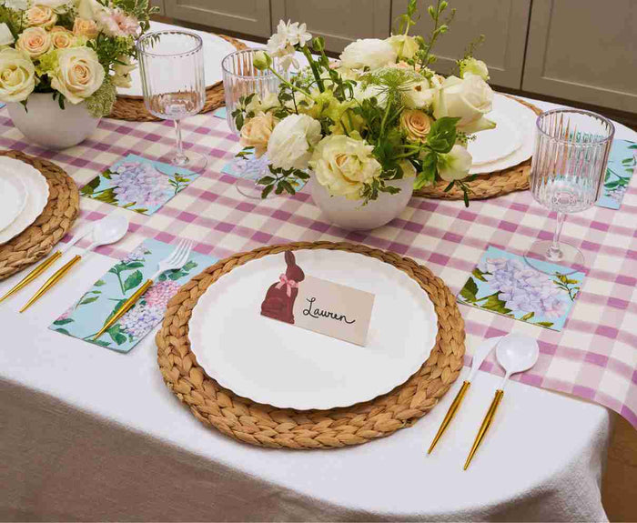 A table set with Sophistiplate’s Ultimate Easter Tablescape for 16 in Violet Hydrangea features designer disposable plates, gold utensils, woven placemats, floral centerpieces, folded napkins with purple flowers, and Lauren's place card with a bunny illustration.
