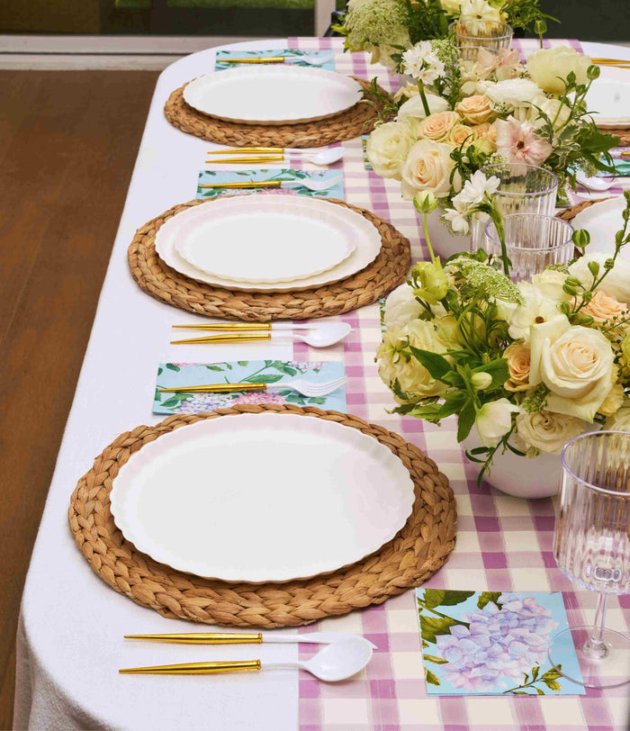 The Ultimate Easter Tablescape for 16: Violet Hydrangea by Sophistiplate LLC features woven placemats, designer disposable plates, gold cutlery, clear glasses, and white and yellow rose floral arrangements on a purple checkered tablecloth.