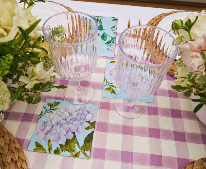 Two clear glasses rest on a purple and white checkered tablecloth, accented by Sophistiplate LLC's Ultimate Easter Tablescape for 16: Violet Hydrangea, with floral-patterned napkins and woven placemats adding charm to the setting.
