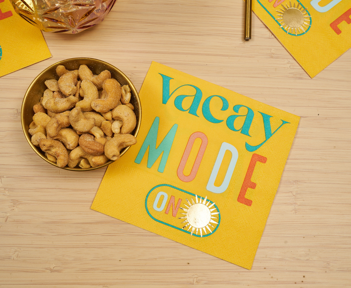 A bowl of cashews sits on a wooden surface beside Sugarplum’s Vacay mode On Cocktail Napkins (26pk), which are bright yellow and feature colorful "vacay MODE ON" text and a small sun illustration.