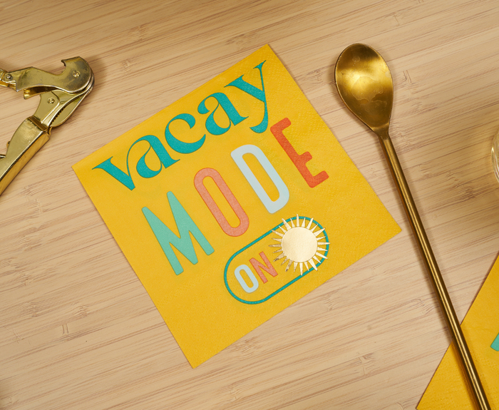 Sugarplum Vacay mode On Cocktail Napkins (26pk) feature colorful "Vacay Mode On" text and a sun illustration on yellow paper, shown on a light wood surface with gold utensils nearby.
