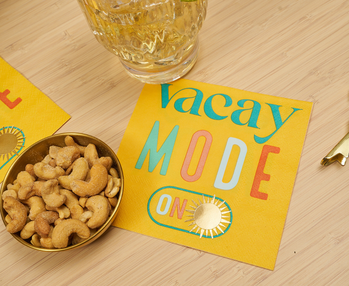 Sugarplum Vacay mode On Cocktail Napkins (26pk) feature colorful “Vacay Mode ON” text on yellow—perfect for summer parties. Shown on a wood table with a drink and cashews.