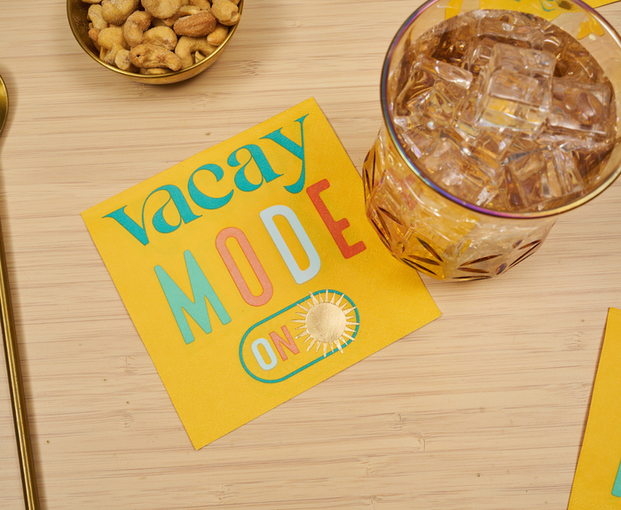 A glass of iced drink and a bowl of peanuts rest on a light wooden surface next to Sugarplum’s Vacay mode On Cocktail Napkins (26pk), featuring vibrant colors, playful summer text, and a cheerful sun illustration.