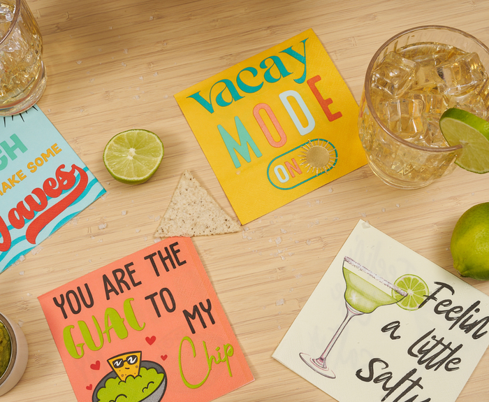 A wooden tabletop displays Sugarplum’s “Feeling a little Salty” Cocktail Napkins (26pk), a lime wedge, tortilla chip, two iced drinks in glasses, and whole limes—perfect for summer entertaining.
