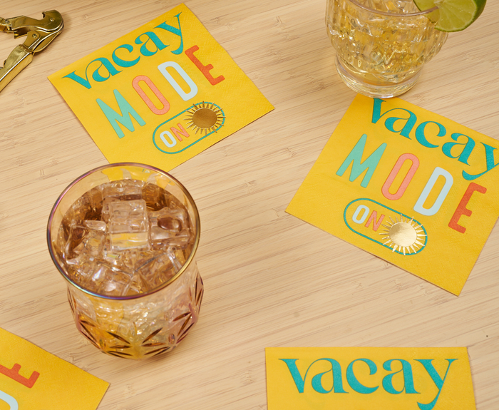 Two glasses with ice and drinks sit on a wooden surface next to Sugarplum’s Vacay mode On Cocktail Napkins (26pk), featuring colorful “Vacay Mode ON” text and a sun graphic. A bottle opener and lime wedge complete the scene.